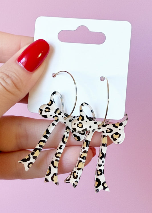 Leopard print earrings on a branded card held by a hand with red nail polish against a pink background.
