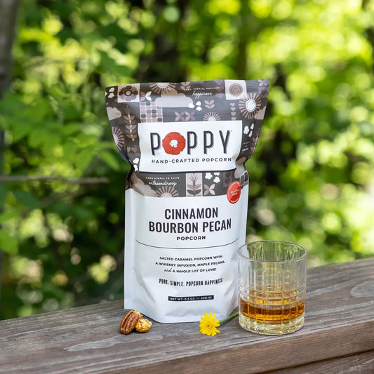 A bag of Poppy Hand-Crafted Popcorn in Cinnamon Bourbon Pecan flavor sits on a wooden railing outdoors, with a glass of amber liquid and two pecans in front of a blurred green forest background.