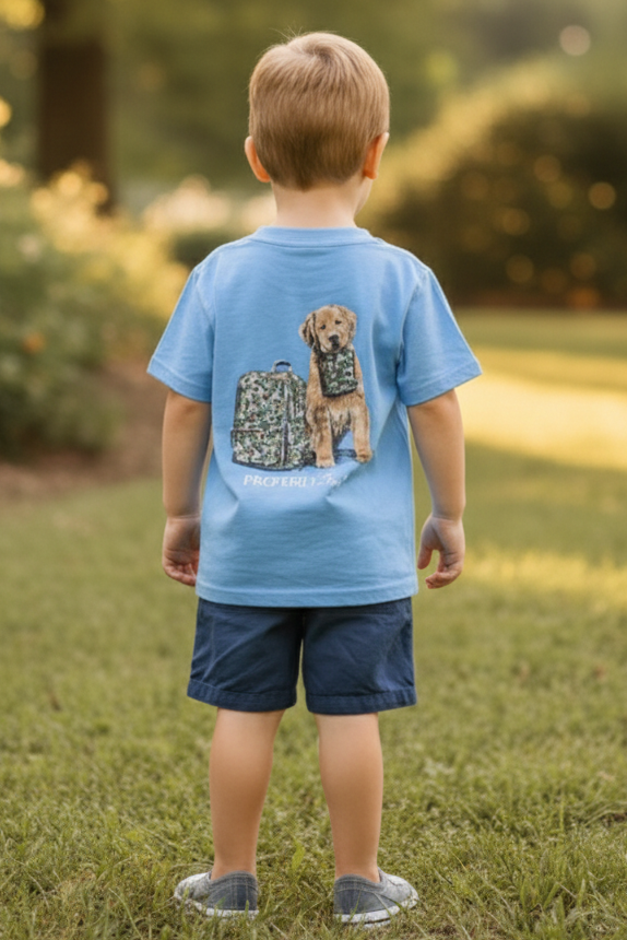 A young child viewed from behind wearing a light blue t-shirt with a graphic of a puppy and suitcase on the back, navy shorts, and gray slip-on shoes, standing on a grassy lawn.