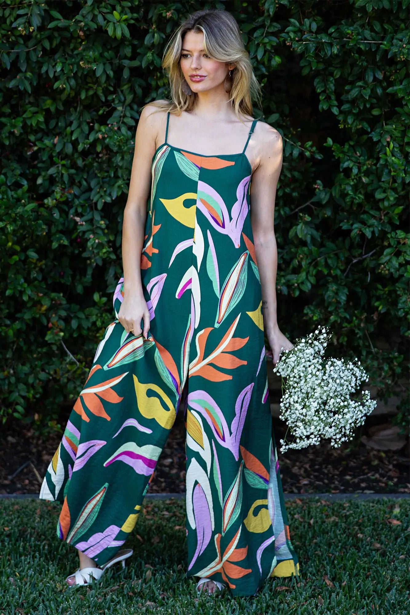 Woman wearing a colorful floral jumpsuit standing outdoors with greenery in the background