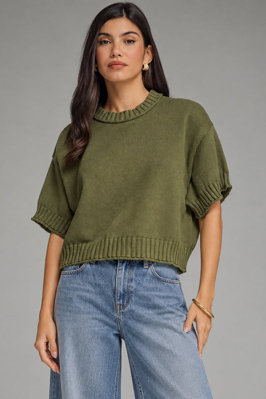 Woman wearing a green sweater and beige pants against a gray background