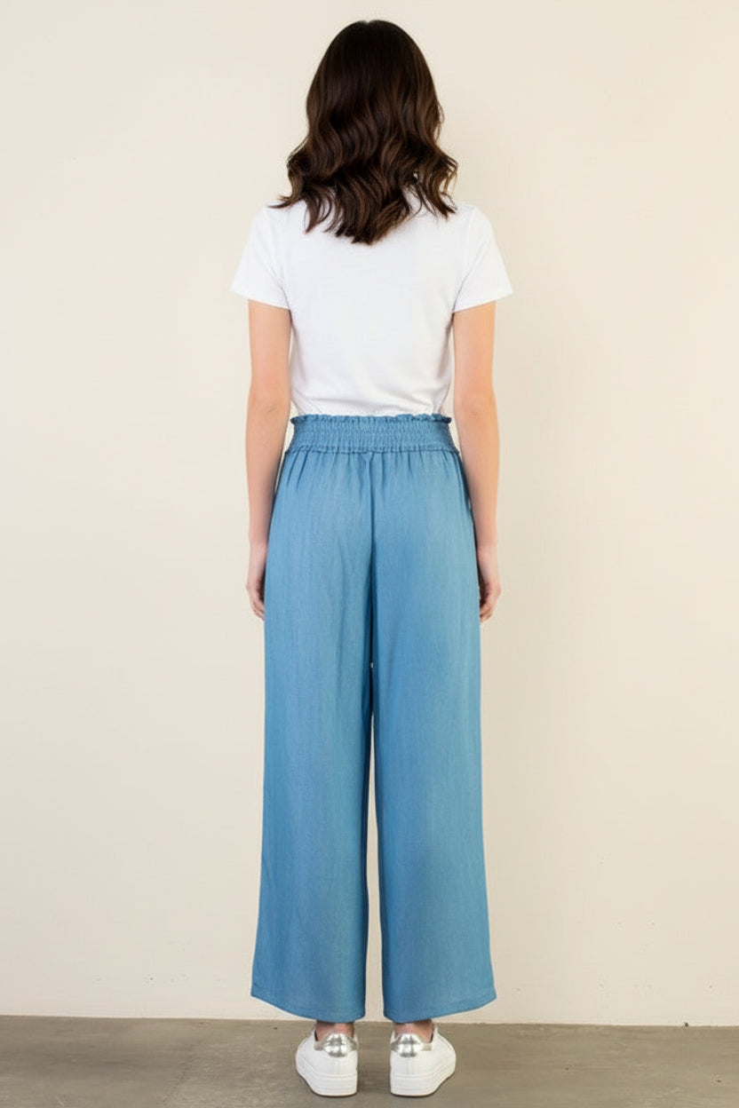 Woman wearing light blue wide-leg pants and a white top on a plain background