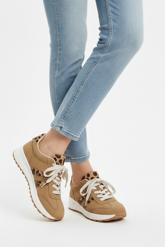 A pair of tan low-top sneakers with leopard-print accents and white laces, shown on a model wearing light-wash blue jeans against a plain white studio background.