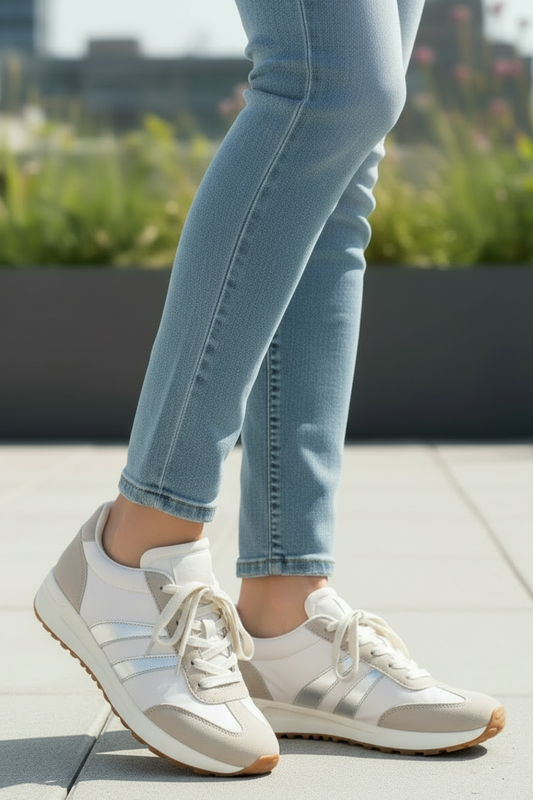 White and beige low-top sneakers with light blue jeans, shown on a person outdoors on a paved surface with blurred greenery in the background.