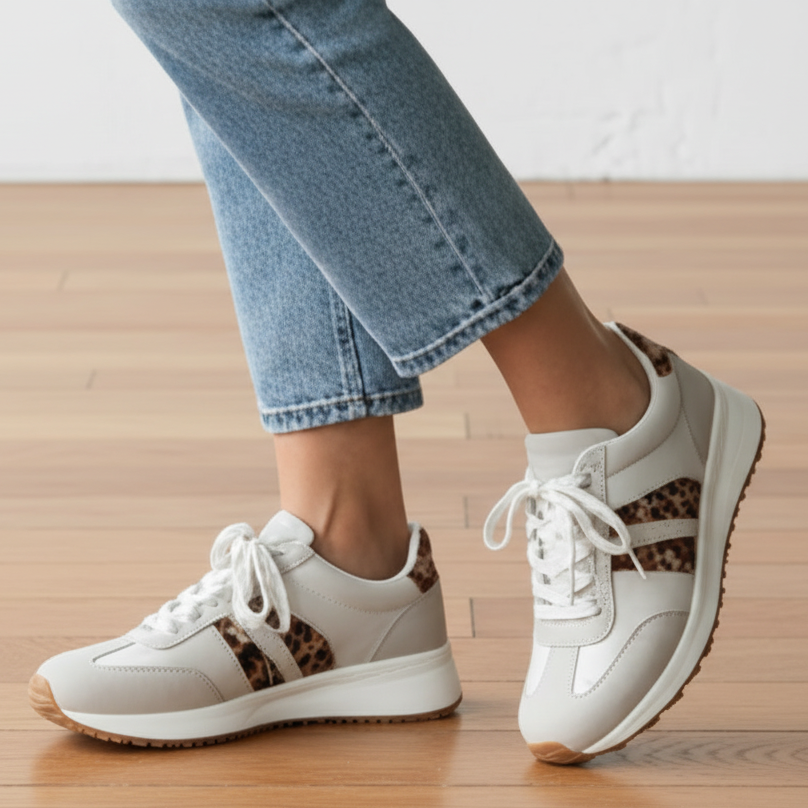 White sneakers with leopard print details worn with blue jeans on a wooden floor.