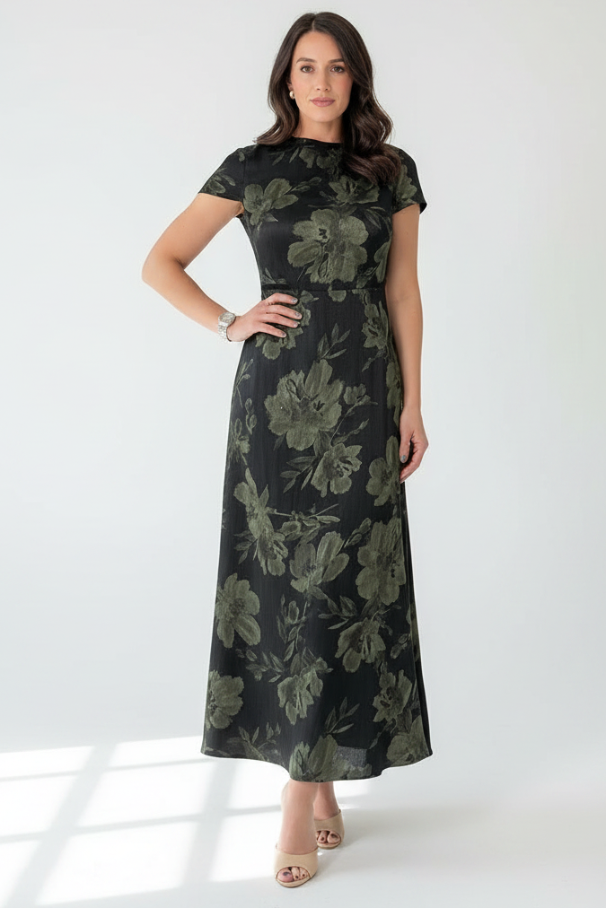 A woman modeling a short-sleeve, high-neck maxi dress with a dark green and black floral pattern, paired with beige heeled sandals, set against a plain white studio background.