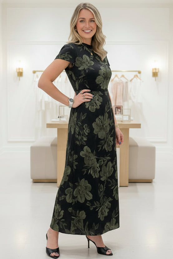 A woman takes a mirror selfie in a bedroom showing a black midi dress with a green floral pattern. The dress has short sleeves, a high neckline, and a fitted waist with a flared skirt.