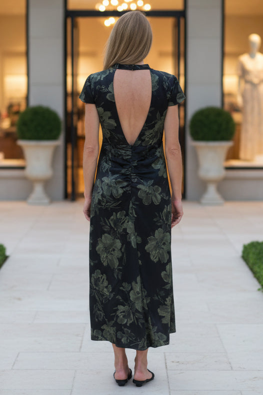 A woman viewed from behind wearing a short-sleeve black dress with a green floral pattern and a deep open back, standing on a concrete path in a backyard with grass, plants, and a house visible.