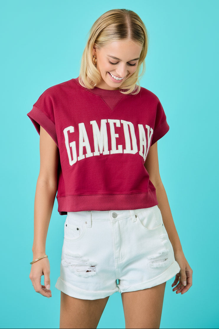 A woman wearing a cropped maroon sweatshirt with 'GAMEDAY' printed in white arched letters across the front, paired with white distressed denim shorts, set against a bright turquoise background.