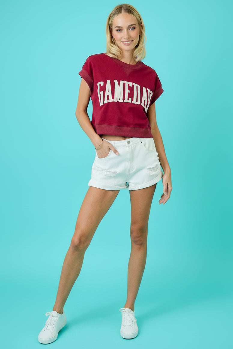 A woman modeling a maroon cropped t-shirt with 'GAMEDAY' printed in white across the chest, paired with white denim shorts and white pointed-toe boots, standing in front of a turquoise background with stacked red, green, and gray milk crates.