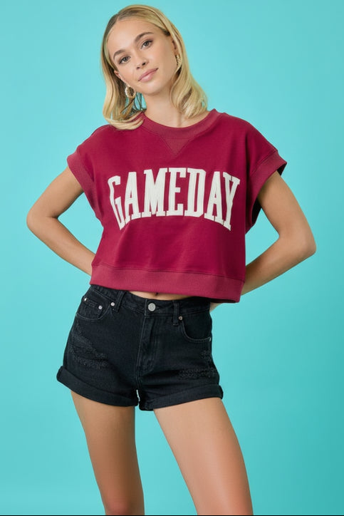 A woman modeling a cropped crewneck sweatshirt in washed burgundy with 'GAMEDAY' printed across the front in white varsity letters, paired with white denim shorts against a solid turquoise background.