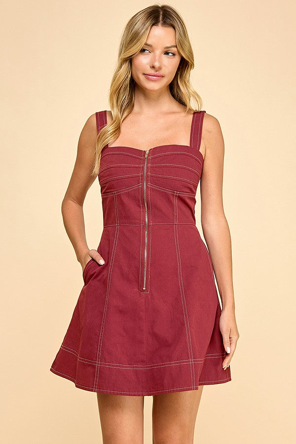 A woman modeling a short, sleeveless A-line dress in a dusty rose or mauve color, featuring a sweetheart neckline, a front zipper, and contrast stitching, set against a solid beige background.