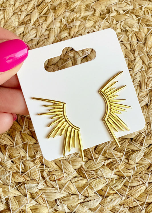 Gold wing-shaped earrings on a white card with 'SR' branding, held over a textured beige surface.