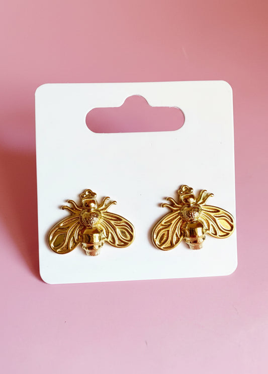 Gold bee-shaped earrings on a white card with 'SR' logo against a pink background