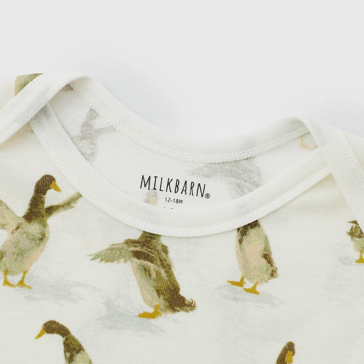 Close-up of a white baby top with a duck print, showing the collar with the brand name MILKBARN.