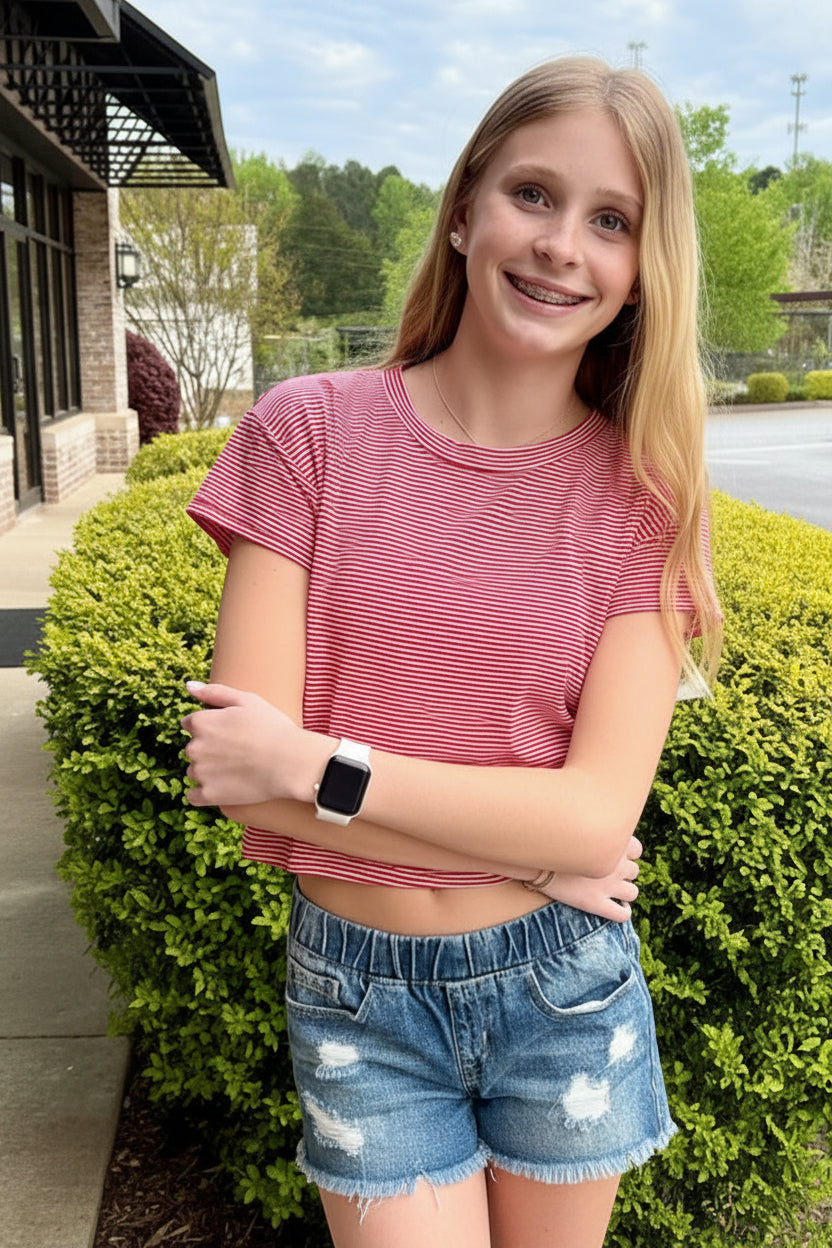 Young girl in a red striped shirt and denim shorts standing outdoors on a sidewalk.