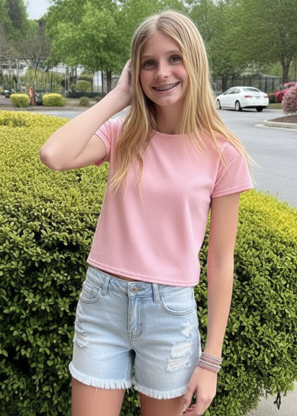 Person in pink shirt and denim shorts standing outdoors with a hedge and vehicle in the background