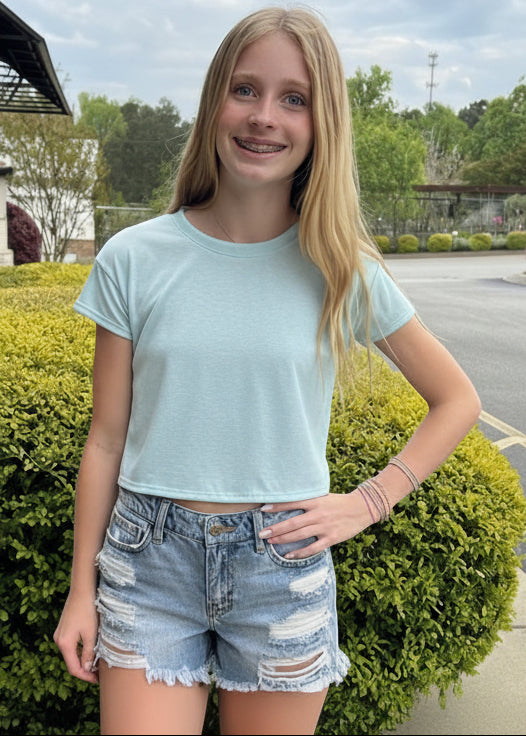 Young girl in light blue t-shirt and denim shorts standing outdoors.