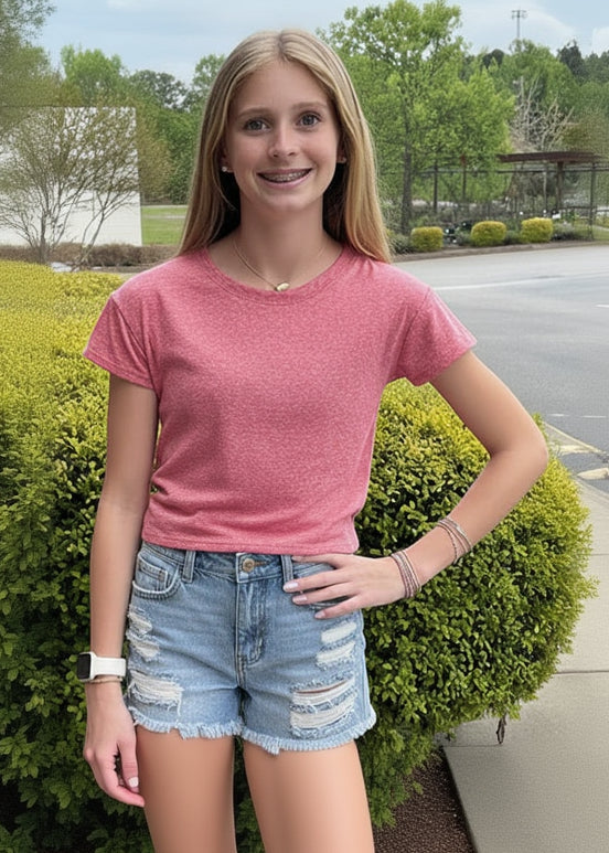 Woman in red shirt and denim shorts standing outdoors with a bush in the background