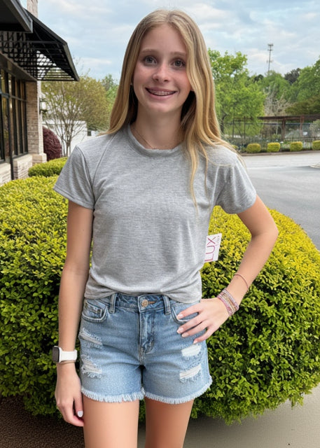 Person wearing a gray t-shirt and denim shorts standing outdoors with greenery and a building in the background.