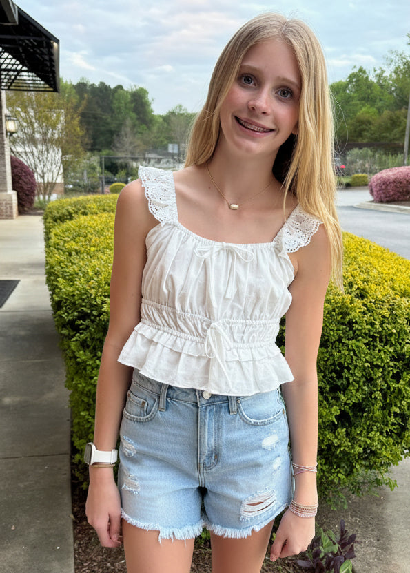 Young woman in a white top and denim shorts sitting outdoors on a concrete ledge.
