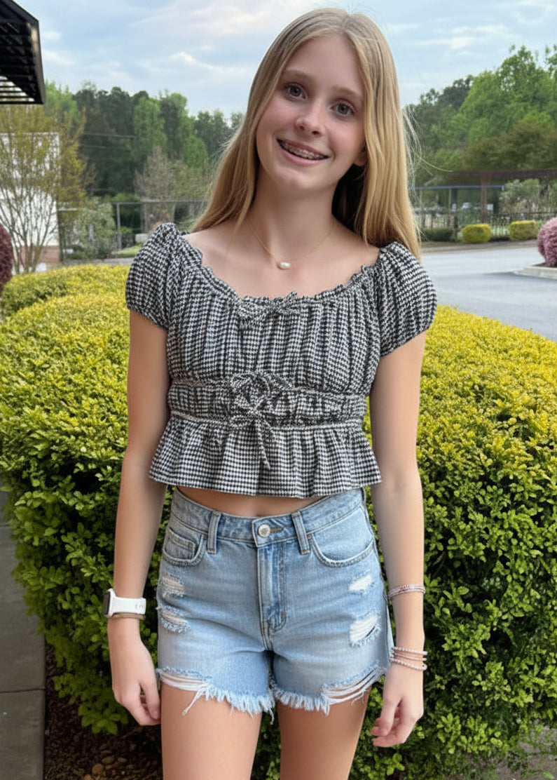 girl in a checkered top and denim shorts standing outdoors near greenery.