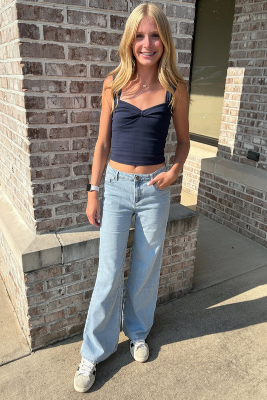 A woman stands outdoors in front of a brick building, wearing a dark navy blue ribbed crop top, light blue wide-leg jeans, and white sneakers.