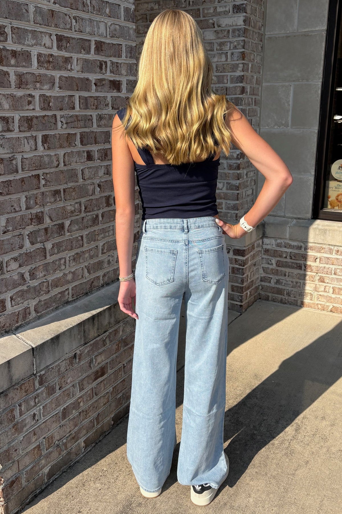 A woman with long blonde hair is shown from behind, wearing a dark navy sleeveless top and light blue wide-leg jeans, standing outdoors on a concrete sidewalk next to a brick building.