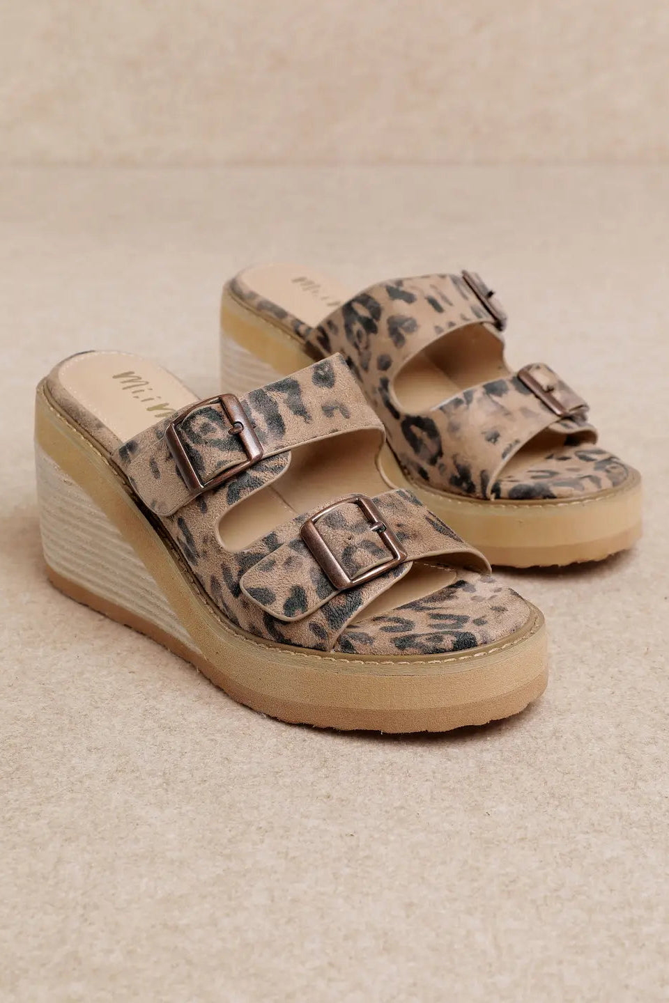 A pair of platform wedge mule sandals featuring a beige and black leopard print upper with double buckle straps, set against a neutral beige background.