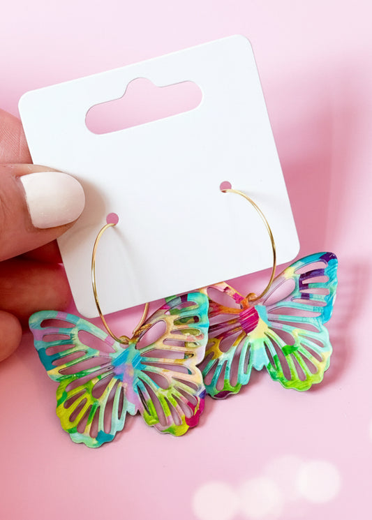 Colorful butterfly-shaped earrings on a SandyRuzzo branded card against a pink background