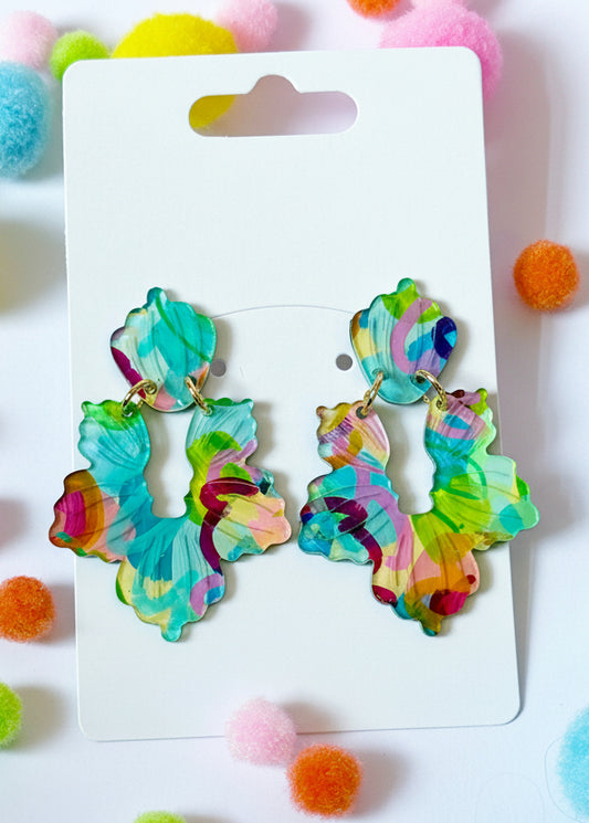 Colorful earrings on a Sandy Rizzo branded card with colorful pom-poms in the background.