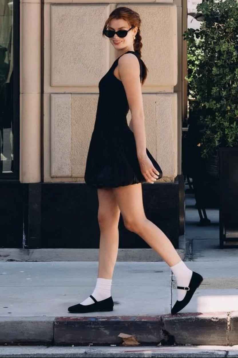 A woman walking on a city sidewalk wearing a sleeveless black mini dress, black loafers, white socks, and sunglasses, with a yellow fire hydrant and beige building facade in the background.