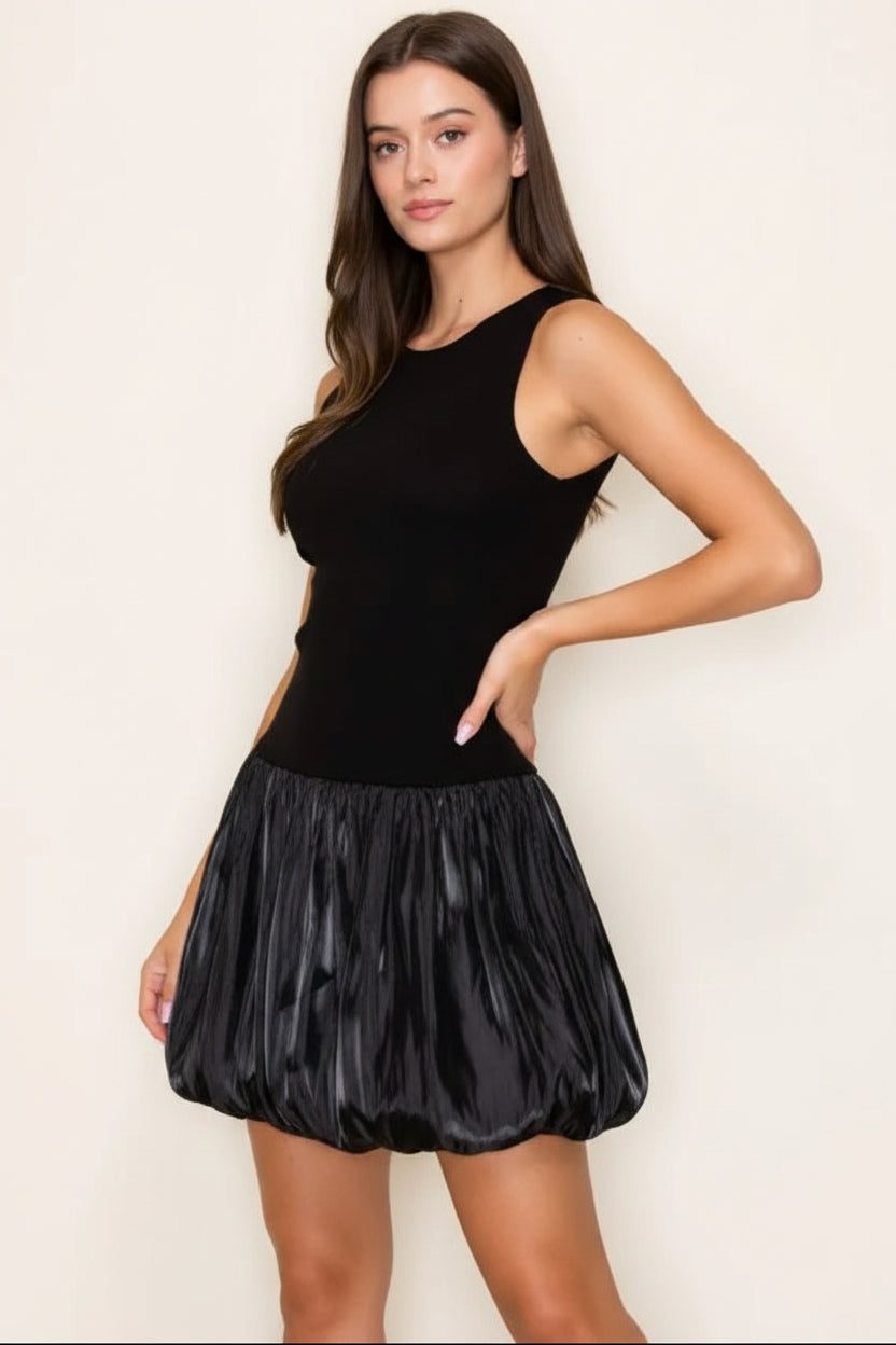 A woman modeling a sleeveless black mini dress with a solid black bodice and a shiny, textured black pleated skirt, set against a plain off-white background.