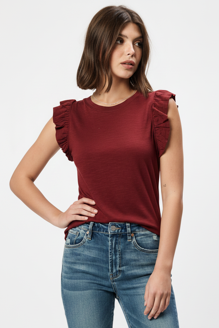 A woman wearing a sleeveless crewneck top in a deep burgundy red, featuring ruffled detailing on the shoulders, paired with blue jeans against a plain white background.