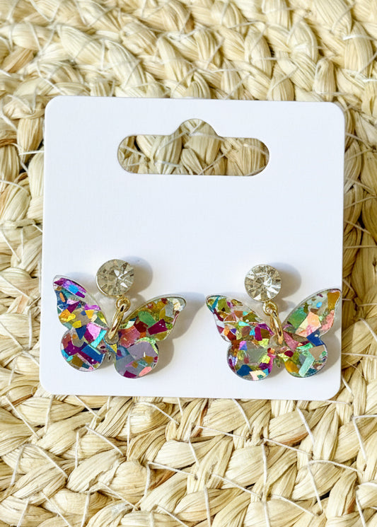 Butterfly-shaped earrings on a branded card with 'SR' logo, placed on a woven mat background.
