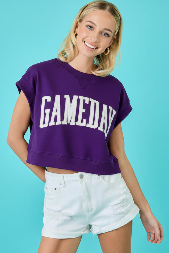 A woman wearing a faded maroon cropped t-shirt with 'GAMEDAY' printed in white across the chest, paired with white denim shorts, set against a bright turquoise background.