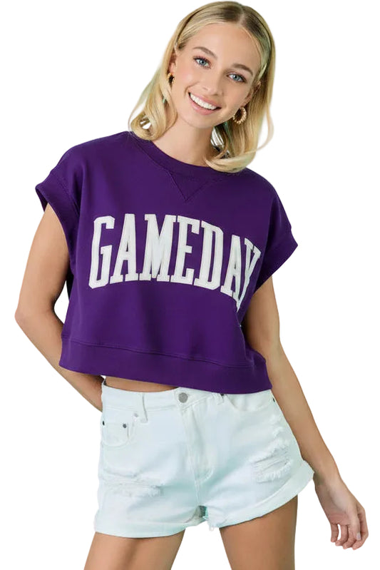 A woman wearing a faded maroon cropped t-shirt with 'GAMEDAY' printed in white across the chest, paired with white denim shorts, set against a bright turquoise background.
