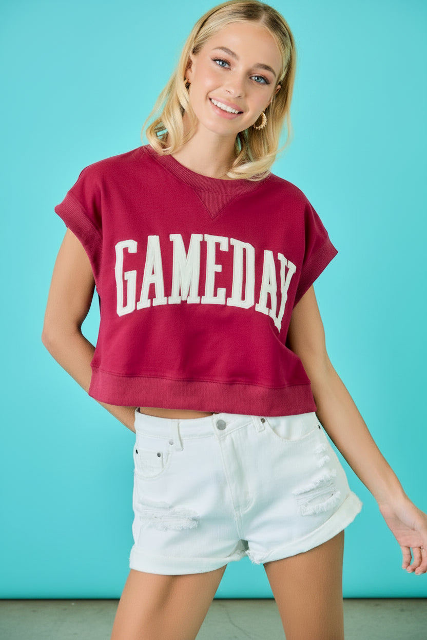 A woman wearing a faded maroon cropped t-shirt with 'GAMEDAY' printed in white across the chest, paired with white denim shorts, set against a bright turquoise background.