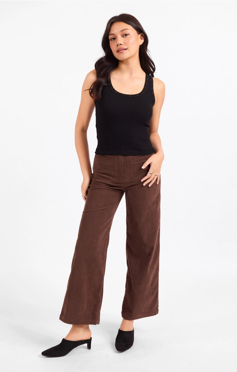 A woman modeling a pair of brown wide-leg cropped pants with a black ribbed tank top and black mule heels, standing against a plain white studio background.