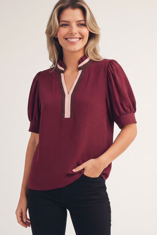 A woman wearing a burgundy short-sleeve blouse with puff sleeves and a split V-neckline trimmed in beige and dark brown, paired with white pants, against a plain light background.