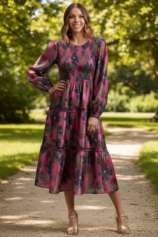 A woman modeling a long-sleeve midi dress with a vibrant pink, purple, and blue abstract floral print, paired with metallic gold wedge sandals, against a solid neutral studio background.