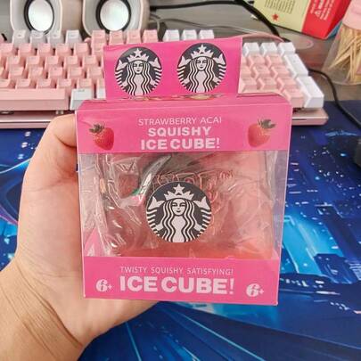 A hand holding a pink Starbucks-branded squishy ice cube toy in its original packaging, with a pink keyboard and desk mat visible in the background.