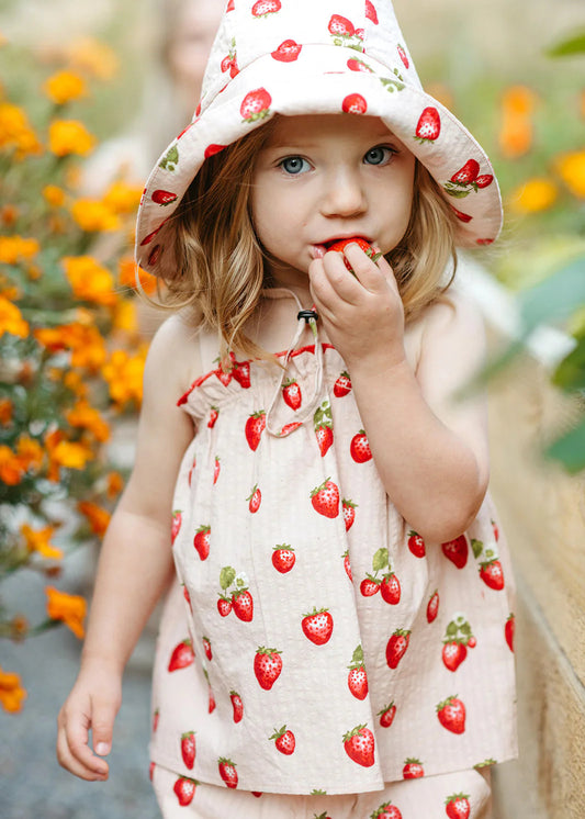 Child wearing a strawberry-patterned outfit and hat, eating a strawberry with a blurred floral background.