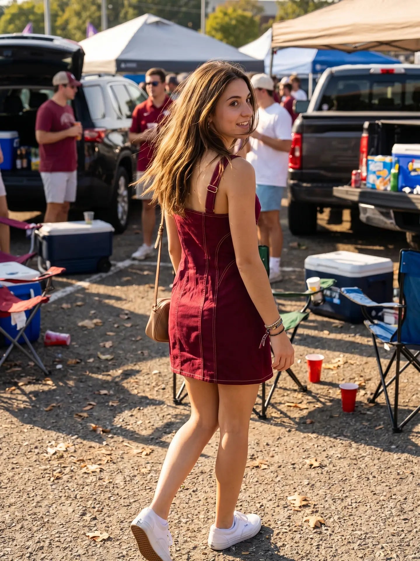 A woman wearing a burgundy corduroy mini dress and white sneakers walks through an outdoor tailgate or festival setting with tents, pickup trucks, and coolers in the background.