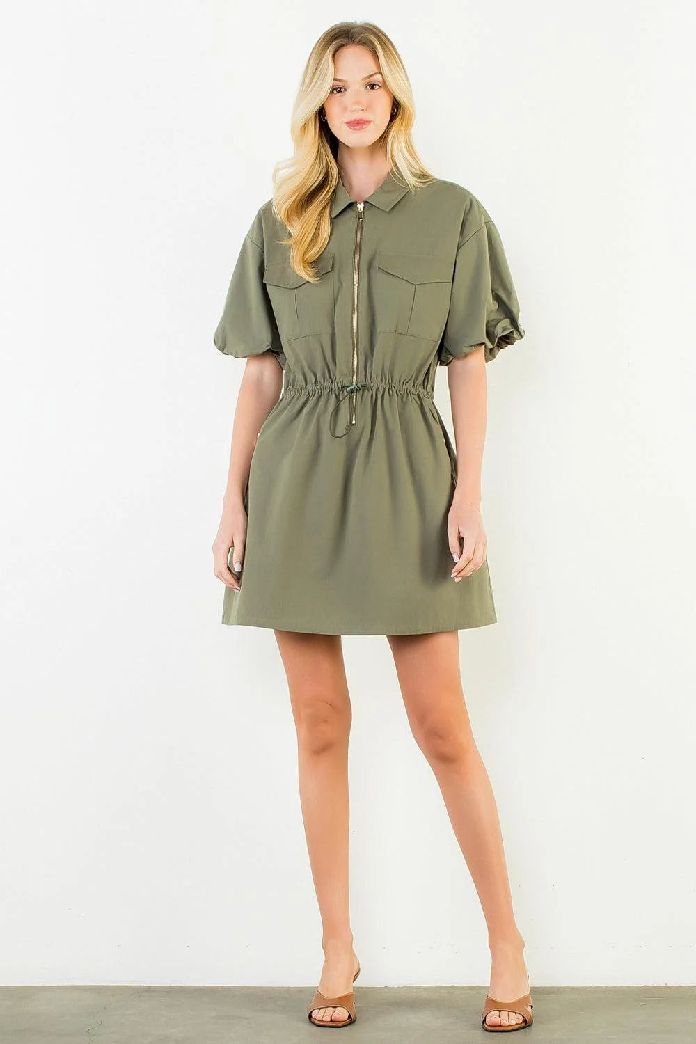 A woman modeling an olive green short-sleeve mini dress with a front zipper, chest pockets, and a drawstring waist, standing against a plain white wall.