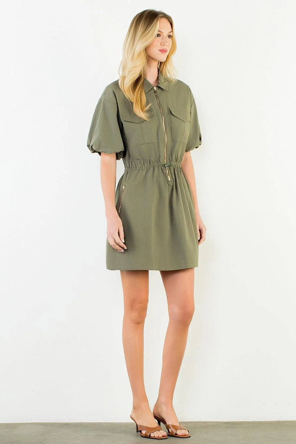 A woman modeling an olive green short-sleeve mini dress with a collared neckline, front zipper, chest pockets, and a drawstring waist, standing against a plain white wall.