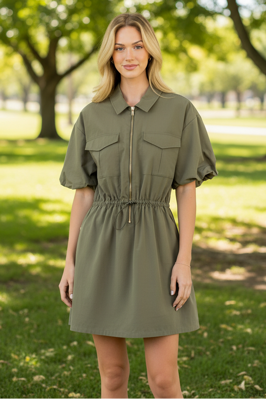 A woman stands outdoors in a park wearing an olive green short-sleeve mini dress with puff sleeves, a collar, front flap pockets, and a drawstring waist.