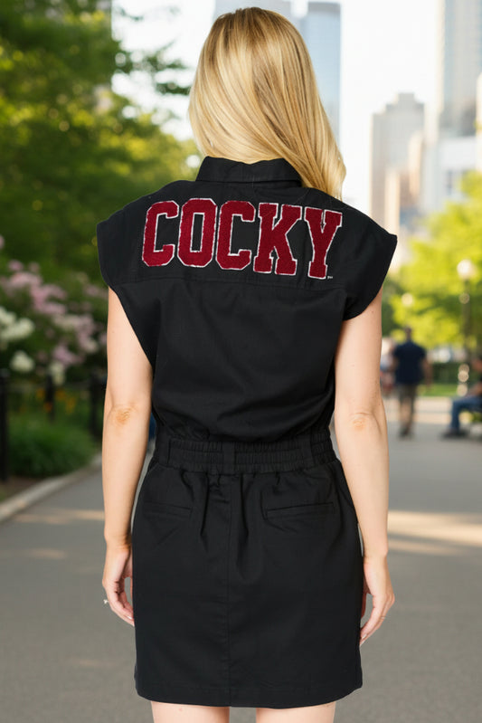 Person wearing a black sleeveless dress with 'COCKY' printed on the back in an urban setting.