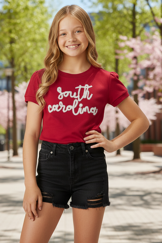 A young girl with blonde hair smiling outdoors, wearing a red short-sleeve t-shirt with 'south carolina' written in white script across the chest, paired with black distressed denim shorts.