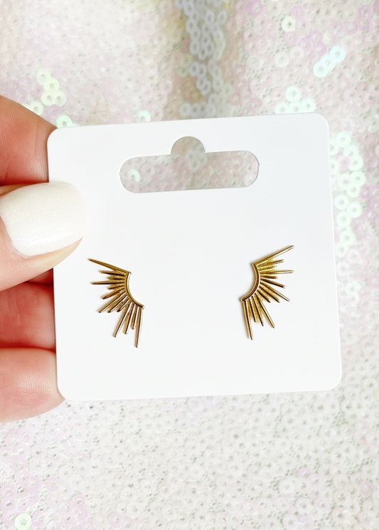 Gold wing-shaped earrings on a Sandy Rizzo branded card held by a hand against a glittery background.